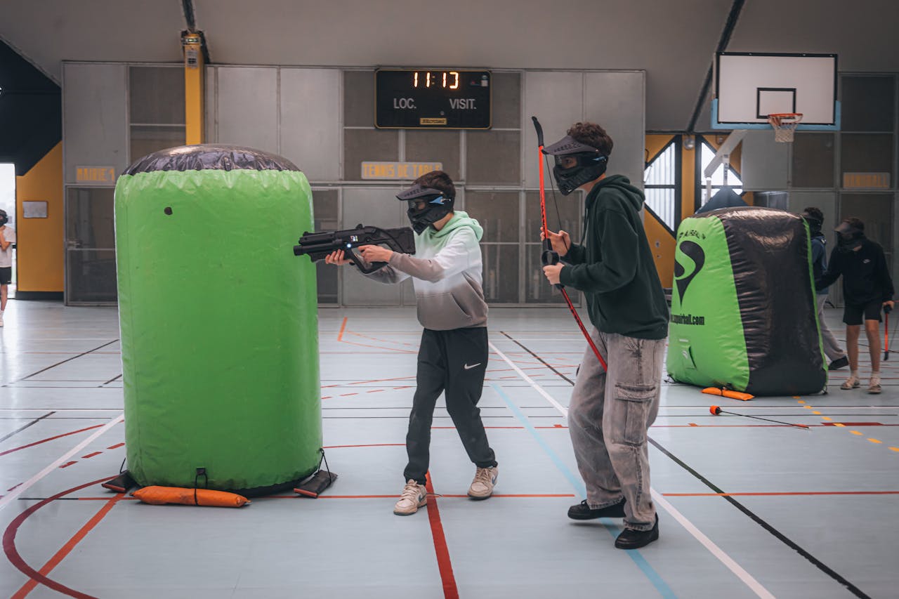 Two people playing indoor paintball wearing masks and using inflatable barriers for cover.